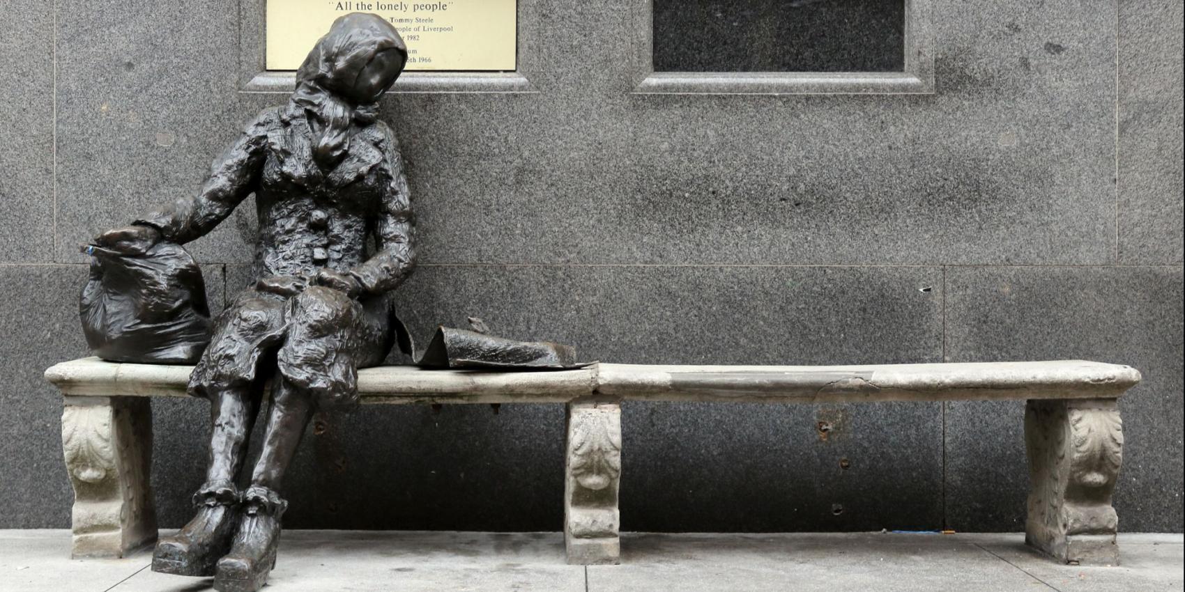 Das Foto zeigt eine Statur aus Stein, die auf einer Bank sitzt vor einer Handwand.