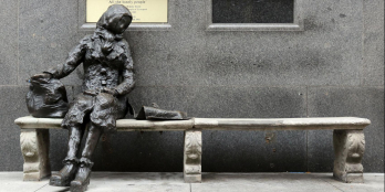 Das Foto zeigt eine Statur aus Stein, die auf einer Bank sitzt vor einer Handwand.