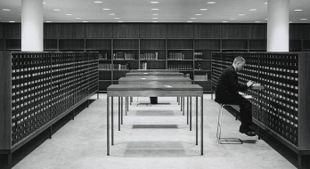 Zu sehen ist ein Bibliotheksraum, in der Mitte Tische, links und rechts Karteischränke. An dem rechten Karteischrank sitzt ein Mann und zieht einen Karteikasten heraus.