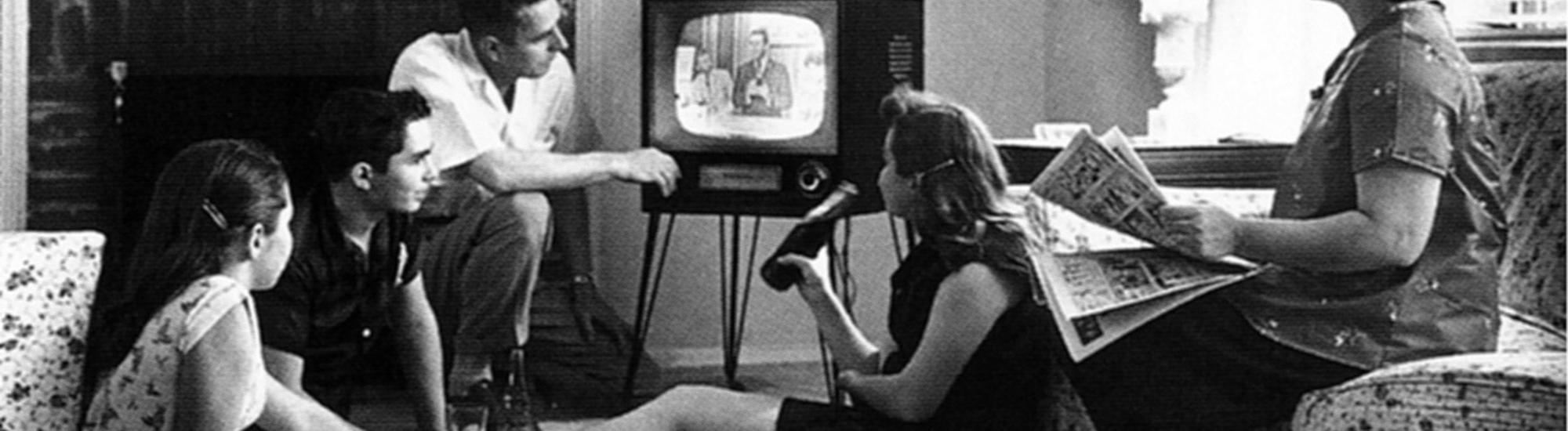 Bild einer amerikanischen Familie beim Fernsehsehen, ca 1958. Fünf Menschen sind um einen Fernseher gruppiert, teilweise liegen oder sitzen sie am Boden, eine Frau sitzt auf einem Sofa und hält eine Zeitung in der Hand. Foto: Evert F. Baumgardner