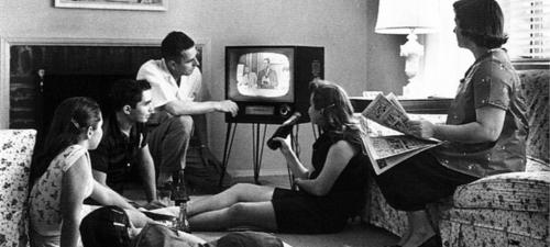 Bild einer amerikanischen Familie beim Fernsehsehen, ca 1958. Fünf Menschen sind um einen Fernseher gruppiert, teilweise liegen oder sitzen sie am Boden, eine Frau sitzt auf einem Sofa und hält eine Zeitung in der Hand. Foto: Evert F. Baumgardner