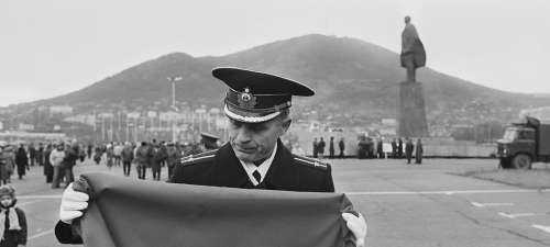 Ein Mann in Militäruniform faltet eine sowjetische Flagge zusammen. Im Hintergrund ein Hügel, davor eine große Statue.